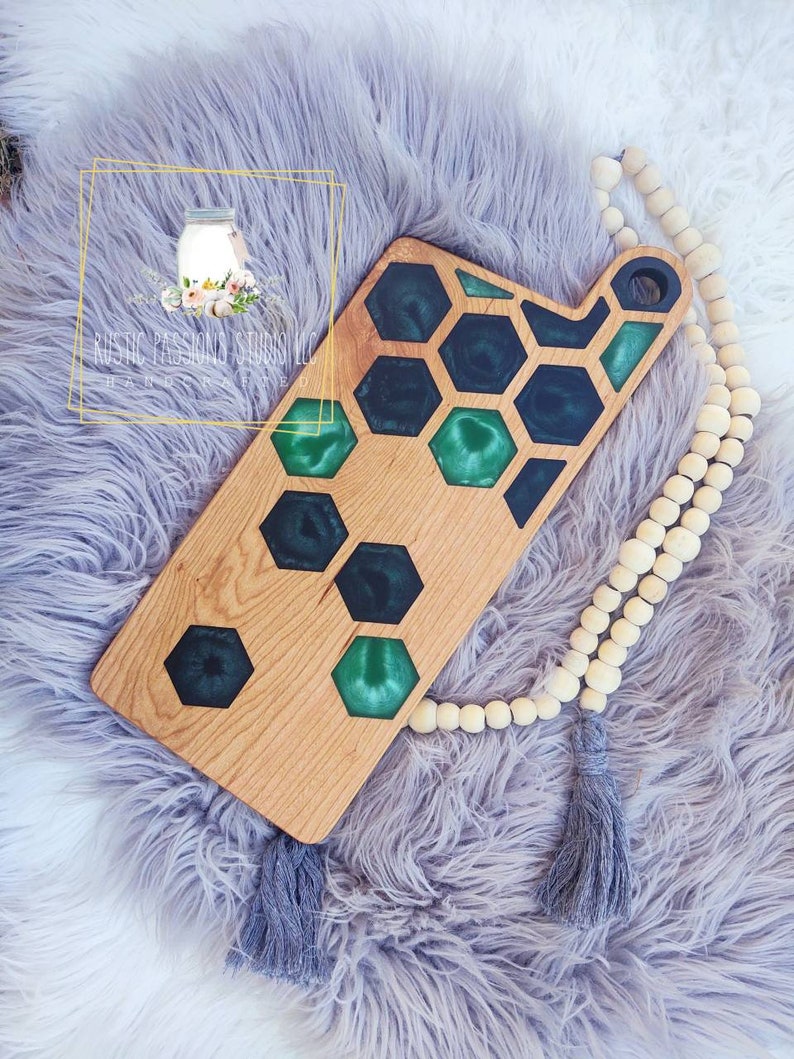 May include: A wooden cutting board with a green and black epoxy resin inlay in a honeycomb pattern. The board has a handle on the side and is handmade.