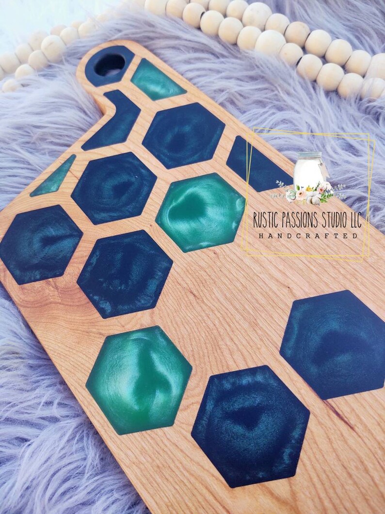 May include: A wooden cutting board with a honeycomb design. The honeycomb is filled with green and blue epoxy resin. The board is handcrafted and has a rustic look.  RUSTIC PASSIONS STUDIO LLC HANDCRAFTED