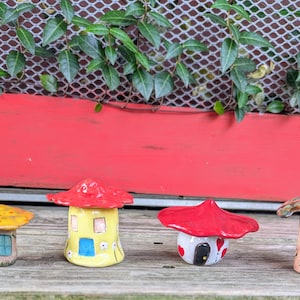 May include: Four ceramic mushroom houses with red, yellow, and brown caps. Each house has a unique design, including doors, windows, and decorative elements. The houses are set against a red background with green foliage.
