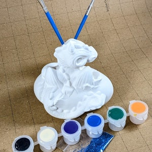 May include: A white ceramic dragon figurine with a paintbrush holder, six small paint pots in black, white, cream, purple, blue, and green, a small bag of blue glitter, and two paintbrushes.