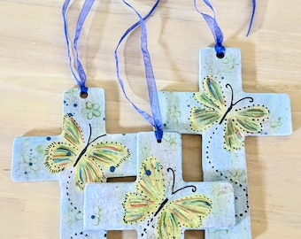 May include: Three ceramic crosses with a blue ribbon. Each cross is painted with a light blue background and a yellow butterfly with black outlines. The butterflies have a speckled pattern of yellow, orange, and green.