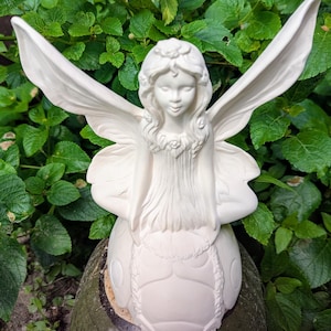 May include: A white ceramic fairy statue with outstretched wings, a floral crown, and a serene expression. The fairy is seated on a rounded base, set against a backdrop of green foliage. This decorative figurine is ideal for garden or home decor.