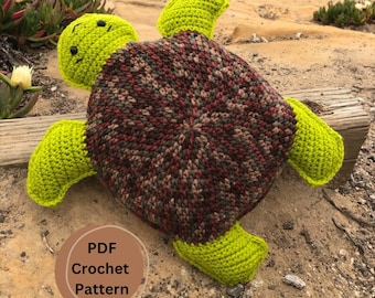 May include: A crocheted sea turtle plush toy with a green shell and brown and red patterned underside. The toy is sitting on a sandy beach with rocks and ocean in the background. The text "PDF Crochet Pattern" is on a brown circle in the lower left corner of the image.
