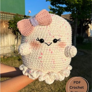 May include: A white crocheted ghost amigurumi with a pink and purple bow, black eyes, and a pink blush. The ghost has a ruffled bottom and is being held in a hand. The text "PDF Crochet Pattern" is in a brown circle on the bottom right corner of the image.