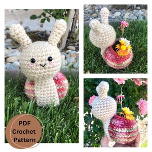 Puede incluir: Un macetero de caracol tejido a crochet con una concha rosa y verde. El caracol tiene un cuerpo blanco y una cara linda. El macetero está lleno de flores pequeñas.