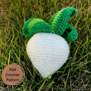 Könnte beinhalten: Eine gehäkelte weiße Rübe mit grünen Blättern. Die Rübe liegt auf einem Bett aus grünem Gras. Das Bild enthält den Text "PDF Crochet Pattern".