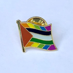 Może przedstawiać: Złota szpilka z wzorem tęczowej flagi. Flaga ma czerwony trójkąt w lewym górnym rogu, po którym następują czarne, białe, zielone, jasnoniebieskie, ciemnoniebieskie i fioletowe pasy.