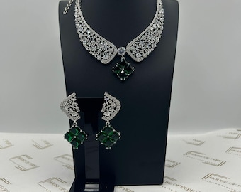 Op de afbeelding: Een zilveren ketting met een groene stenen hanger en bijpassende oorbellen. De ketting is een kraag-stijl met een groot, ingewikkeld ontwerp. De oorbellen zijn hangende stijl met een groene steen aan de onderkant.