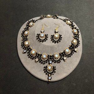 May include: A black and gold beaded necklace and matching earrings set with iridescent stones. The necklace has a pendant drop and the earrings are teardrop shaped.