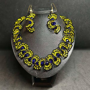 May include: A beaded jewelry set featuring a necklace and matching earrings. The necklace and earrings are made with yellow and black beads, with blue accents. The jewelry is displayed on a black mannequin head.