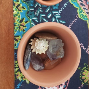 May include: Two terracotta pots filled with various crystals and seashells. The pots are on a blue and green patterned fabric.