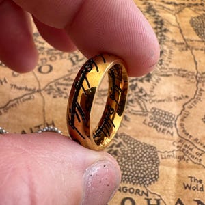 May include: A gold ring with black engraved lettering, held between fingers. The ring's surface reflects light, and the text appears to be a script. The background features a map with place names.