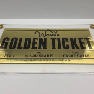May include: A gold-coloured "Wonka Golden Ticket" with black text, encased in clear acrylic and secured with brass-coloured screws. The ticket reads "GOLDEN TICKET" and includes the date "FEB.1" and time "10 A.M.(SHARP)".