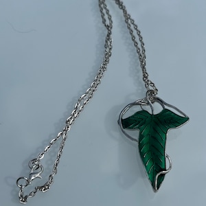 Può includere: Una collana con catena d'argento con un ciondolo a forma di foglia in smalto verde. La foglia è dettagliata con venature e ha uno stelo attorcigliato.