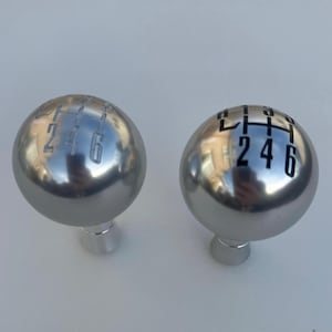 以下が含まれることがあります： 黒い数字と文字が書かれた銀色のシフトノブが2つ。ノブの数字は1、2、4、5、6です。