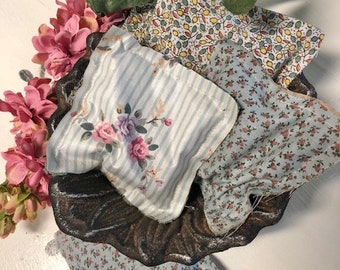 May include: Four small fabric sachets with floral patterns. The sachets are arranged on a dark brown metal tray. The tray has a scalloped edge. The sachets are made of light blue and white fabric with floral prints. The tray is surrounded by pink flowers.