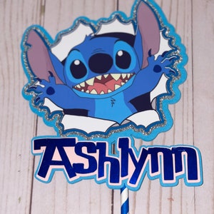 Może przedstawiać: Niebiesko-biały topper na tort z postacią Stitch z filmu Lilo & Stitch, z imieniem Ashlynn napisanym niebieskimi literami.