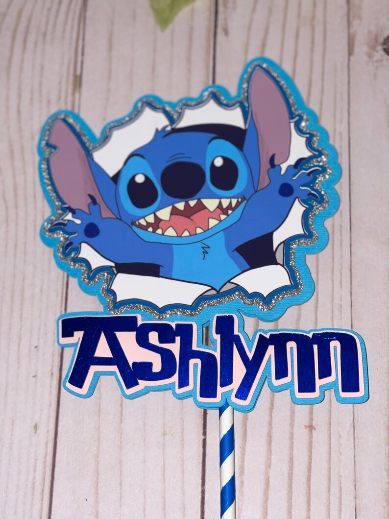 Może przedstawiać: Niebiesko-biały topper na tort z postacią Stitch z filmu Lilo & Stitch, z imieniem Ashlynn napisanym niebieskimi literami.