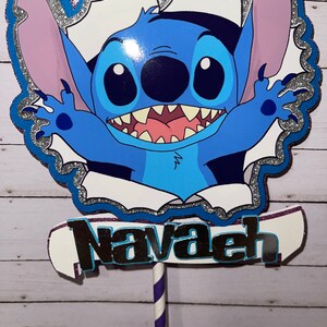 Może przedstawiać: Topper na tort z niebieskim Stitch'em, srebrną błyszczącą ramką i imieniem "Navaeh" napisanym srebrnymi błyszczącymi literami.