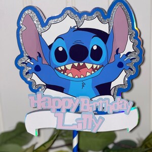 Może przedstawiać: Niebieski topper do tortu z postacią Stitch z białym banerem z napisem "Happy Birthday Lily". Postać otoczona jest srebrną błyszczącą ramką.