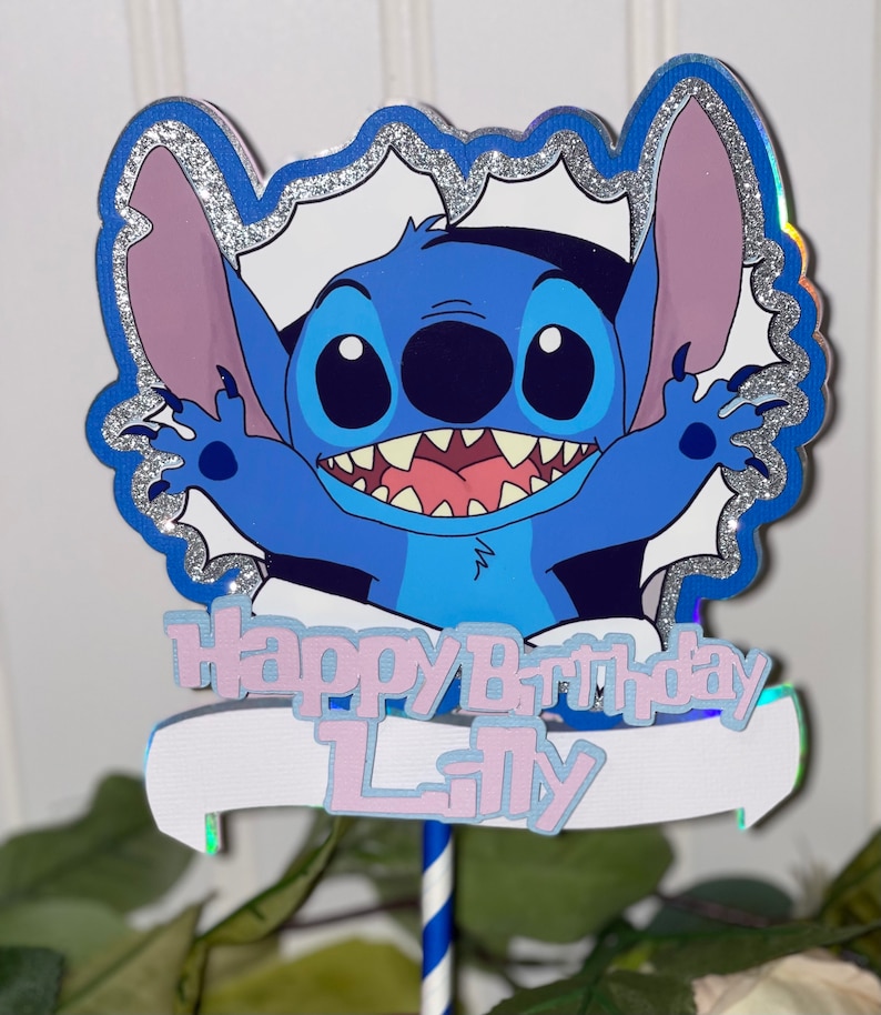 Może przedstawiać: Niebieski topper do tortu z postacią Stitch z białym banerem z napisem "Happy Birthday Lily". Postać otoczona jest srebrną błyszczącą ramką.