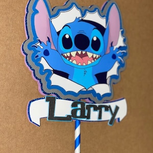 Może przedstawiać: Niebieski i fioletowy topper na tort z postacią z kresk&oacute;wki o dużych oczach i szerokim uśmiechu. Postać nazywa się Stitch i trzyma baner z napisem "Larry".