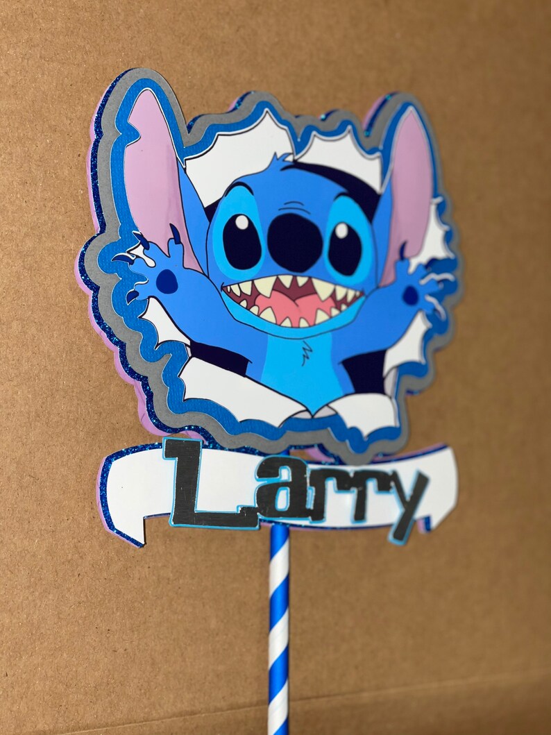 Może przedstawiać: Niebieski i fioletowy topper na tort z postacią z kresk&oacute;wki o dużych oczach i szerokim uśmiechu. Postać nazywa się Stitch i trzyma baner z napisem "Larry".