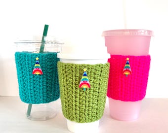 May include: Three crocheted cup sleeves in teal, green, and hot pink, each adorned with a colorful Christmas tree-shaped embellishment. The sleeves are designed to fit around various cup sizes, offering a festive touch.
