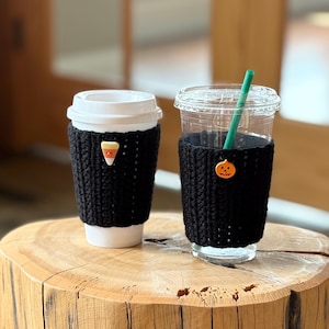 May include: Two coffee cups with black knitted cozies. One cozy has a candy corn button, the other a pumpkin button. A green straw is in the clear cup. The cups are on a wooden surface.