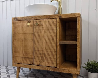 May include: A wooden bathroom vanity with a white vessel sink and gold faucet. The vanity has two doors, a side shelf, and gold knobs. The wood has a natural finish. The vanity sits on angled legs.