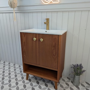 May include: A wooden bathroom vanity unit with a white countertop and a gold tap. The unit has two doors with gold handles and a lower shelf. The floor has a grey and white geometric pattern. A hanging plant and a neon sign are in the background.