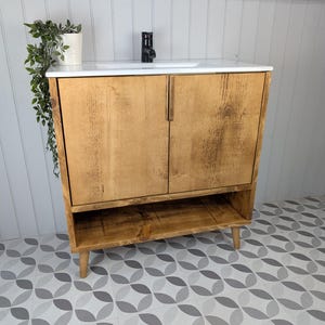 May include: A wooden bathroom vanity unit with a white countertop and a black tap. The cabinet has two doors and a lower shelf. The wood has a natural finish. A white pot with a green plant is on the left. The floor has a grey and white geometric pattern.