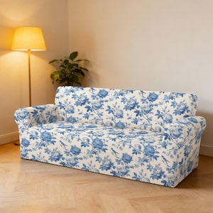 May include: A two-seater sofa with a floral pattern in blue and white. The sofa has a classic design with rolled arms and a skirted base. The floral pattern features roses and other flowers. The sofa is in a well-lit room with a floor lamp and a potted plant.