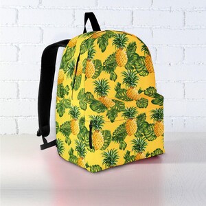 May include: A yellow backpack with a repeating pattern of pineapples and green leaves. The backpack has black straps and a black top handle. The background is a white brick wall and a white surface.