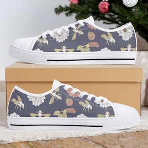 Op de afbeelding: Paar canvas sneakers met witte zolen met een herhalend patroon van bijen, bloemen en aardbeien op een donkerblauwe achtergrond. De sneakers hebben witte veters en een witte neus. De schoenen worden op een kartonnen doos getoond.