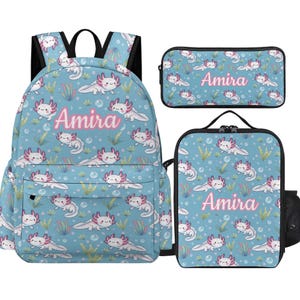 Könnte beinhalten: Ein Rucksack, ein rechteckiges Federmäppchen und eine Lunchtasche, alle mit hellblauem Hintergrund und einem wiederkehrenden Muster aus weißen und rosa Axolotln, grünem Seegras und Blasen. Jeder Artikel zeigt den Namen Amira in rosa Schrift.