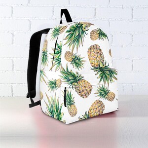 May include: White backpack with a repeating pattern of watercolor pineapples in shades of yellow, green, and brown. The backpack has black straps and a black zipper.