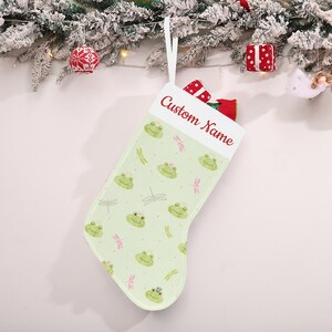 May include: A light green Christmas stocking with a pattern of smiling frog faces wearing crowns and pink dragonflies. The top of the stocking has the text "Custom Name" in red. A white ribbon is attached for hanging.