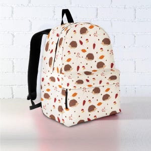 Może przedstawiać: Kremowy plecak z powtarzającym się wzorem brązowych jeży, pomarańczowych i czerwonych liści oraz małych grzybów. Plecak ma czarne paski i czarny zamek błyskawiczny.