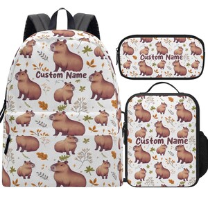 May include: A white backpack, pencil case, and lunch bag set featuring a repeating pattern of brown capybaras and botanical elements. The text "Custom Name" is displayed on each item. The backpack has black straps.