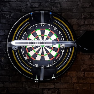 May include: A black dartboard with yellow and white rings around the outside. A dart is stuck in the board, and the numbers 18, 4, 13, 10, 15, 2, 17, 3, 19, 7, 16, 8, 9, 12, 5, 20, 7, 1 are visible.