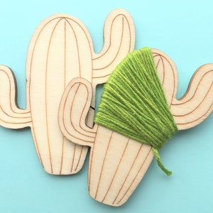May include: Wooden cactus-shaped craft pieces with etched details. One cactus has a bundle of green yarn wrapped around it, suggesting a DIY project or decorative element. The background is a solid light blue.