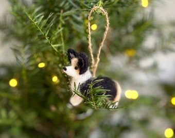 May include: A felt ornament of a black and white dog with brown accents hanging from a tree branch. The ornament is made of wool felt and has a rustic twine hanger.