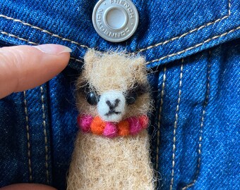 Könnte beinhalten: Ein kleines, braunes Filz-Alpaka mit einer pinkfarbenen und orangenen Perlenkette. Das Alpaka sitzt in einer blauen Jeans-Tasche.