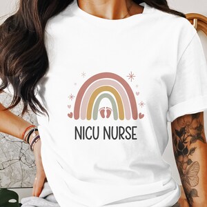 Puede incluir: Camiseta blanca con un diseño de arcoíris y el texto "NICU NURSE". El arcoíris tiene un esquema de color rosa claro, naranja claro, amarillo, verde claro y azul claro. Hay pequeños corazones rosas y estrellas blancas alrededor del arcoíris.