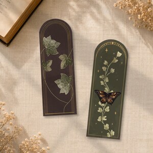 Peut inclure: Deux marque-pages rectangulaires à sommet arrondi. Le marque-page de gauche est marron foncé avec des feuilles de lierre dessinées en doré. Celui de droite est vert foncé avec une arche dorée, une vigne centrale et un papillon marron.
