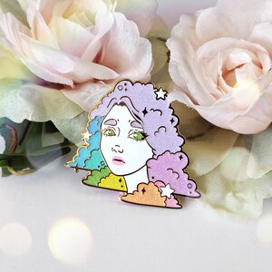 May include: A decorative enamel pin with a face design. The pin features a woman's face with green eyes, pink lips, and multi-colored hair. The hair is a mix of purple, blue, and rainbow colors, with star accents. The pin has a gold outline.