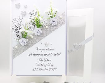 May include: A white wedding card with silver glitter and a silver and white floral design. The card reads "Congratulations Arianna & Harold On Your Wedding Day 27th October 2028".