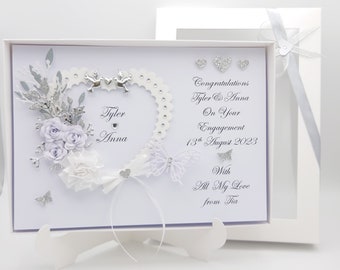 May include: A white engagement card with a heart-shaped design, adorned with silver embellishments, flowers, and a butterfly. The card includes the names "Tyler & Anna" and the date "13th August 2023", with a message of congratulations.