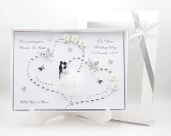 May include: A white wedding card with a silhouette of a bride and groom, adorned with rhinestones, butterflies, and white flowers. The card is presented in a white box with a silver ribbon. Text reads "Congratulations Charles & Steph" and "On Your Wedding Day 4th November 2023".